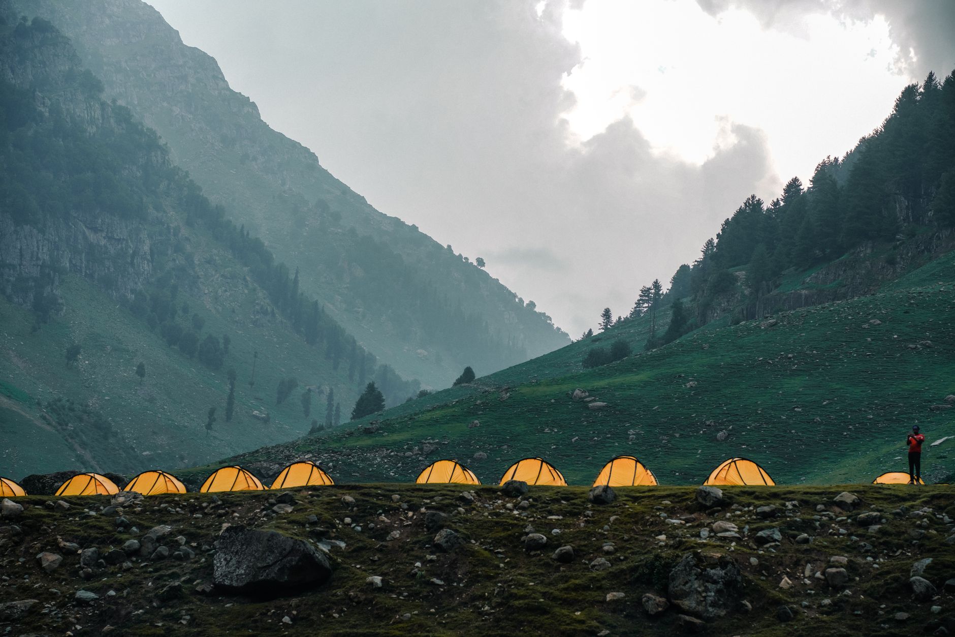 Scenic mountain campsite with illuminated tents and lone hiker under cloudy sky.