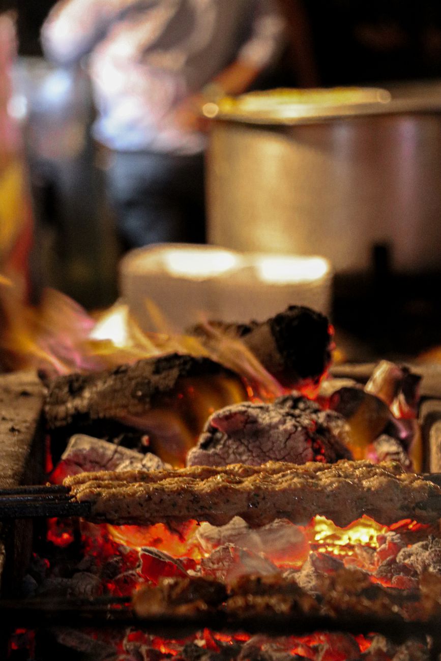 A close-up shot of sizzling meat grilled over wood fire flames, creating a rustic cooking scene.